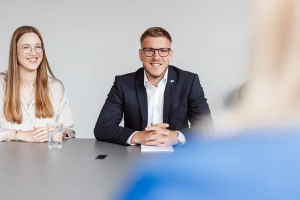 Zwei Personen sitzen an einem Tisch, gegenüber von einer dritten Person, von der man nur den Hinterkopf sieht. Eine Person trägt ein helles Hemd, die andere ein dunkles Sakko.