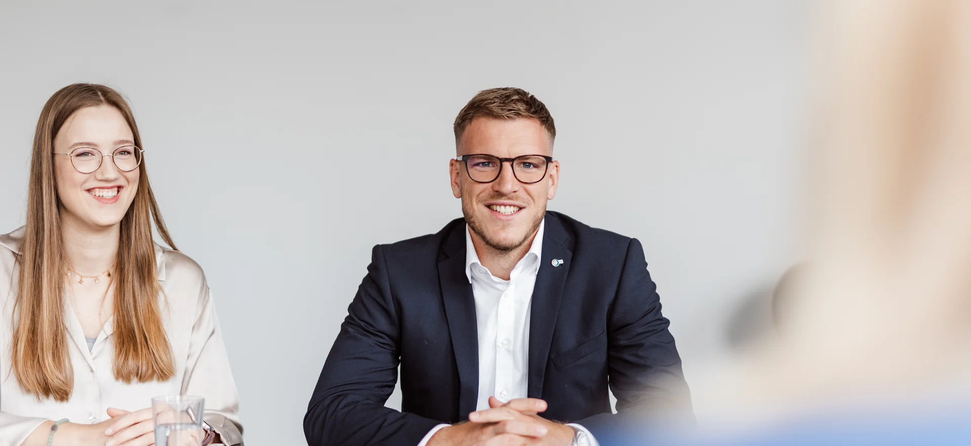 Zwei Personen sitzen an einem Tisch, gegenüber von einer dritten Person, von der man nur den Hinterkopf sieht. Eine Person trägt ein helles Hemd, die andere ein dunkles Sakko.