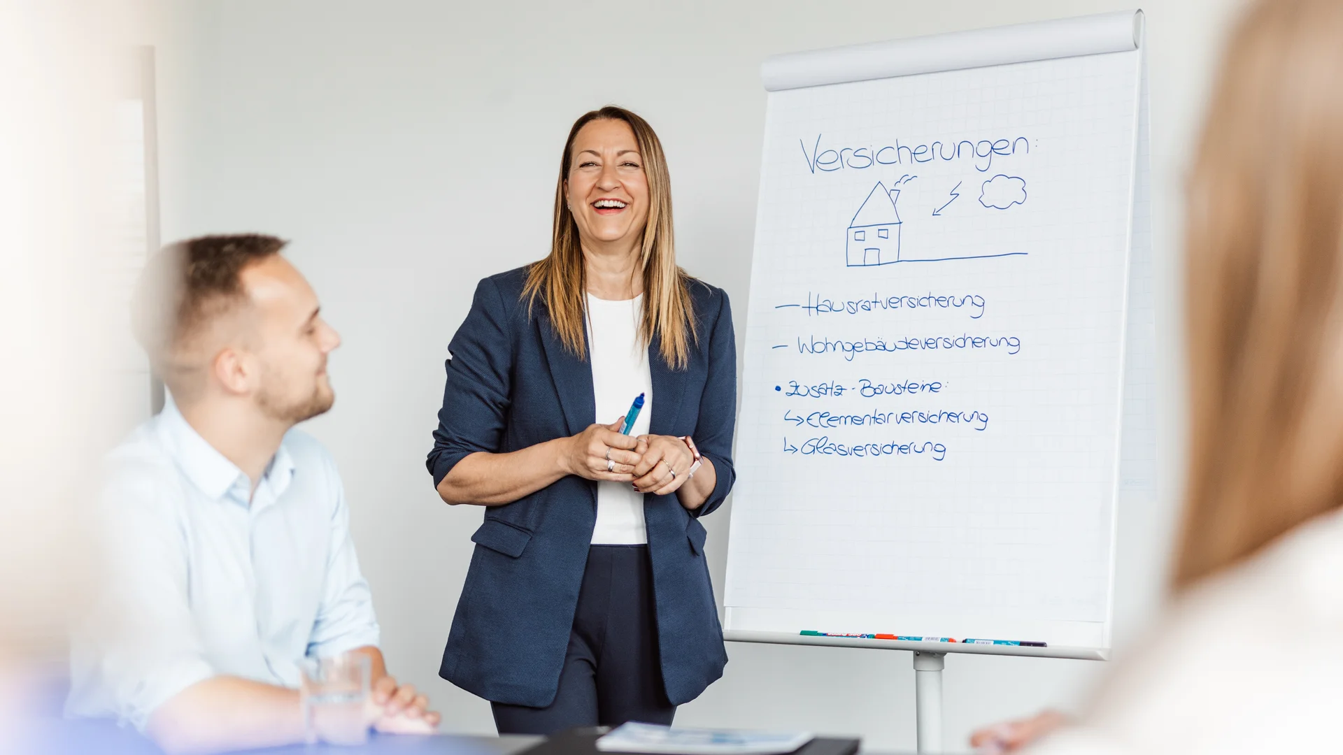 Frau in blauer Busineskleidung steht vor Flipchart, hat Stift in der hand und lacht in die Kamera
