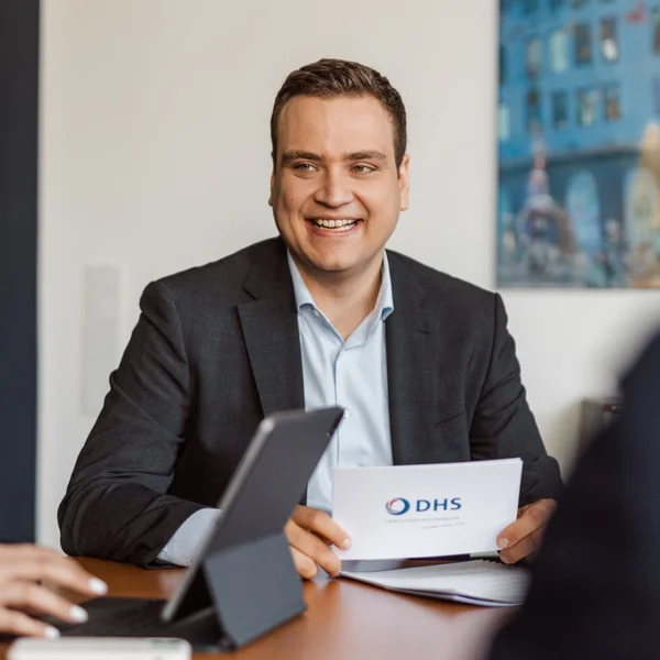 Fünf Personen in Businesskleidung sitzen an einem Konferenztisch, ein Mann hält ein Blatt mit dem Logo 'DHS' und lächelt