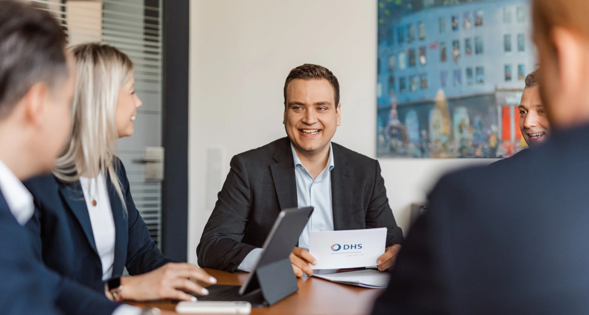 Fünf Personen in Businesskleidung sitzen an einem Konferenztisch, ein Mann hält ein Blatt mit dem Logo 'DHS' und lächelt