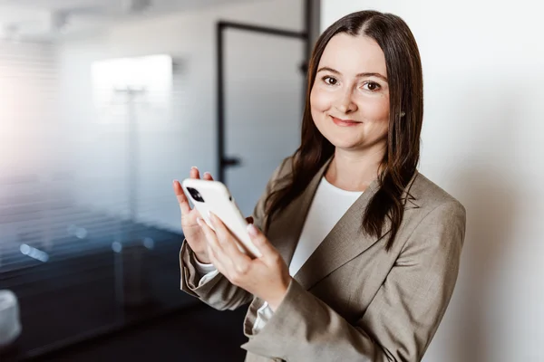 Lächelnde Frau mit dunklen Haaren in hellbraunem Blazer hält ein Smartphone in den Händen