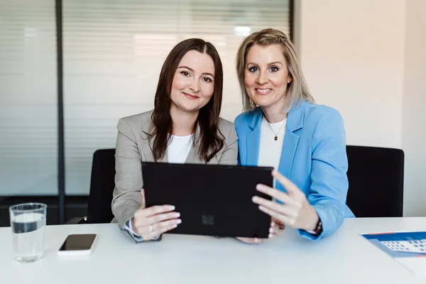 Brünette Frau in beige farbenem Blazer und blonde Frau in blauem Blazer sitzen gemeinsam vor laptop, beide lächeln in Kamera