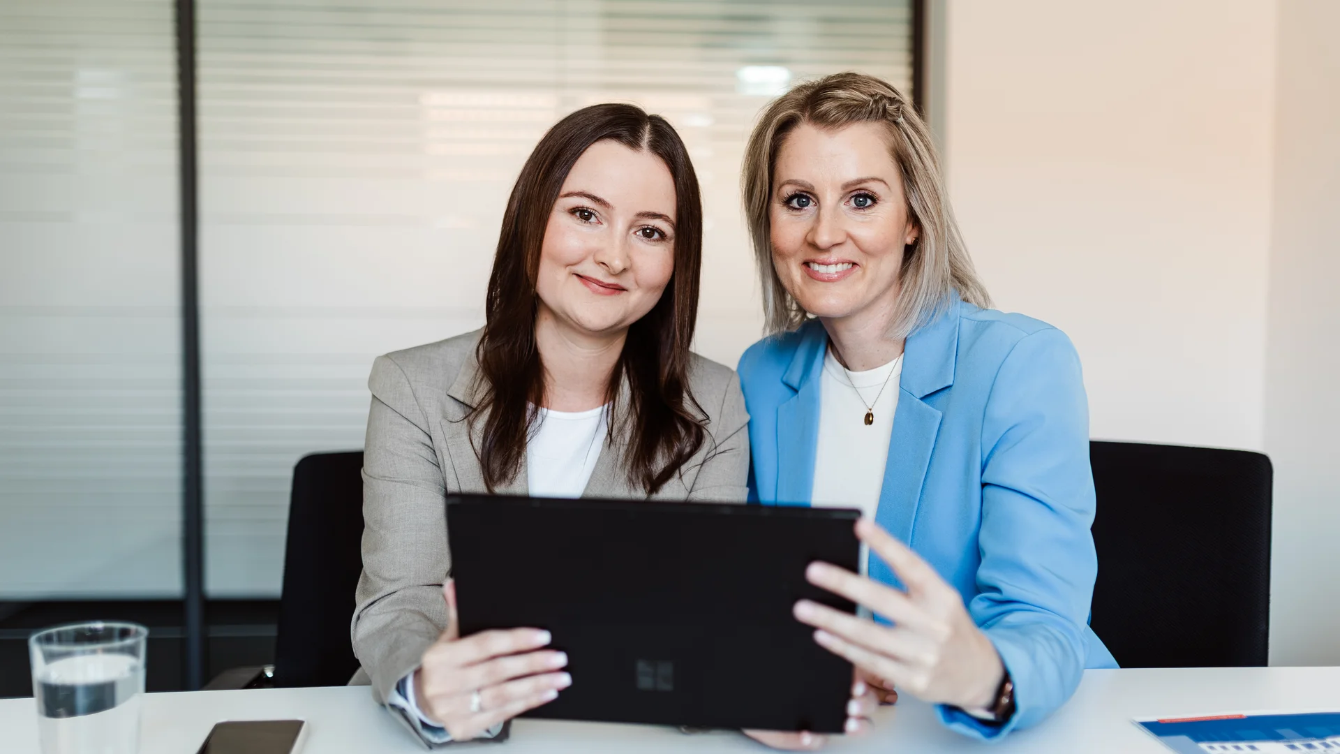 Brünette Frau in beige farbenem Blazer und blonde Frau in blauem Blazer sitzen gemeinsam vor laptop, beide lächeln in Kamera