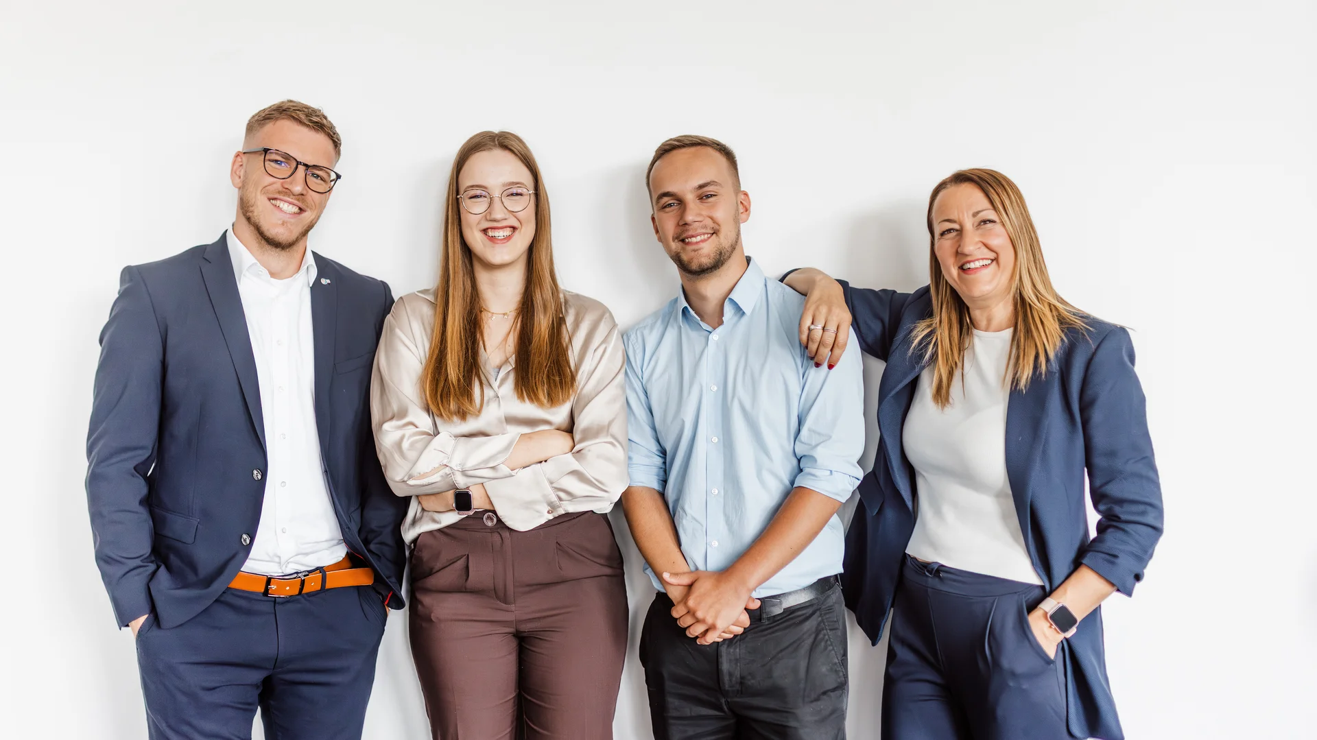 Zwei Männer und zwei Frauen in Businesskleidung stehen nebeneinander, wirken vertraut und schauen in die Kamera