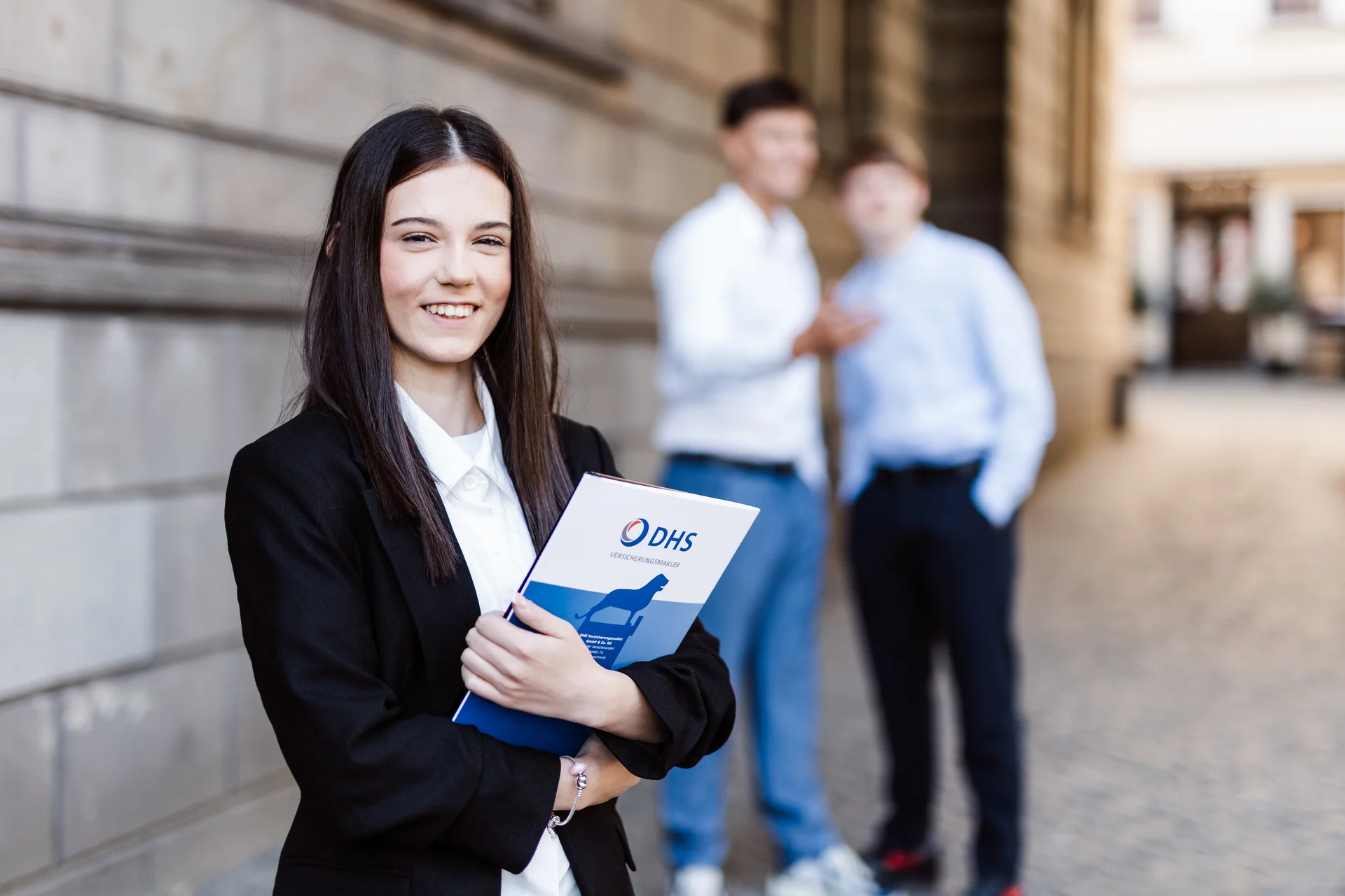 Eine Auszubildene mit dunklem Haar und dunklem Sacko steht mit einer Broschüre in der Hand vor einem Gebäude und lächelt in die Kamera