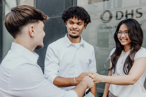 Drei junge Menschen in weißer Kleidung begrüßen sich freundlich mit Handschlag vor einem Gebäude mit dem Schriftzug 'DHS Versicherungsmakler'