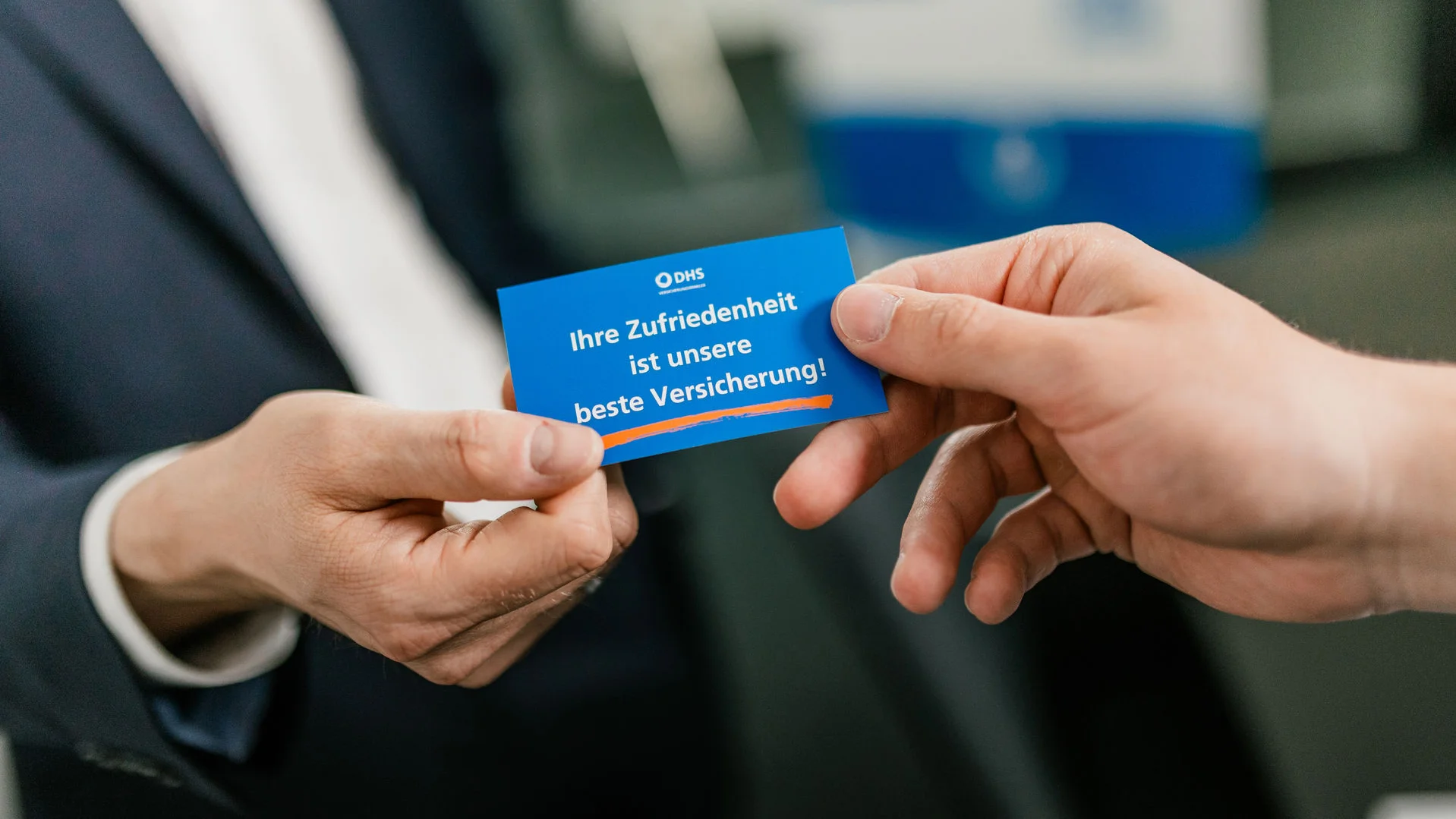 Man sieht zwei Hände, die eine gibt der anderen eine Karte im Visitenkartenformat wo folgender Text drauf steht: Ihre Zufriedenheit ist unsere beste Versicherung