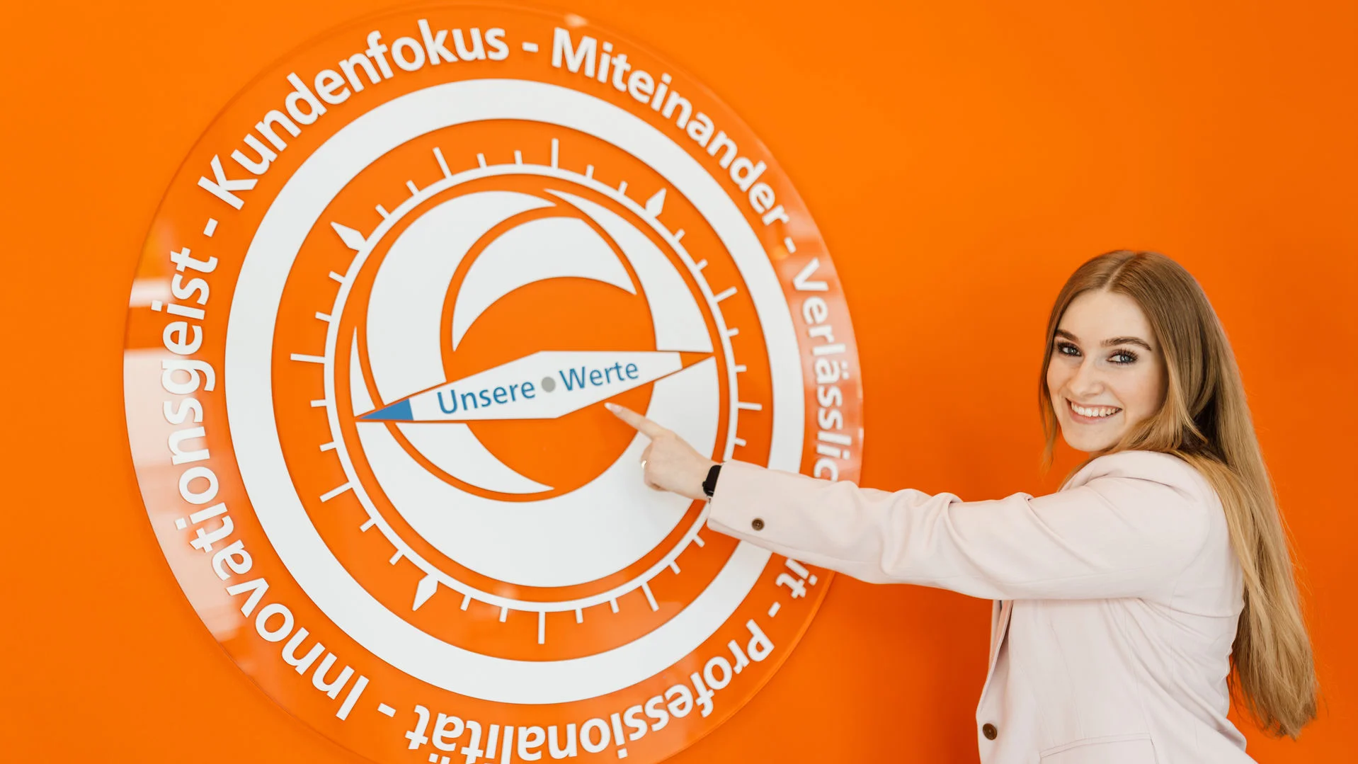 Kompass mit Unternehmenswerten auf orange farbener Wand, junge Frau in Businesskleidung steht daneben und zeigt darauf