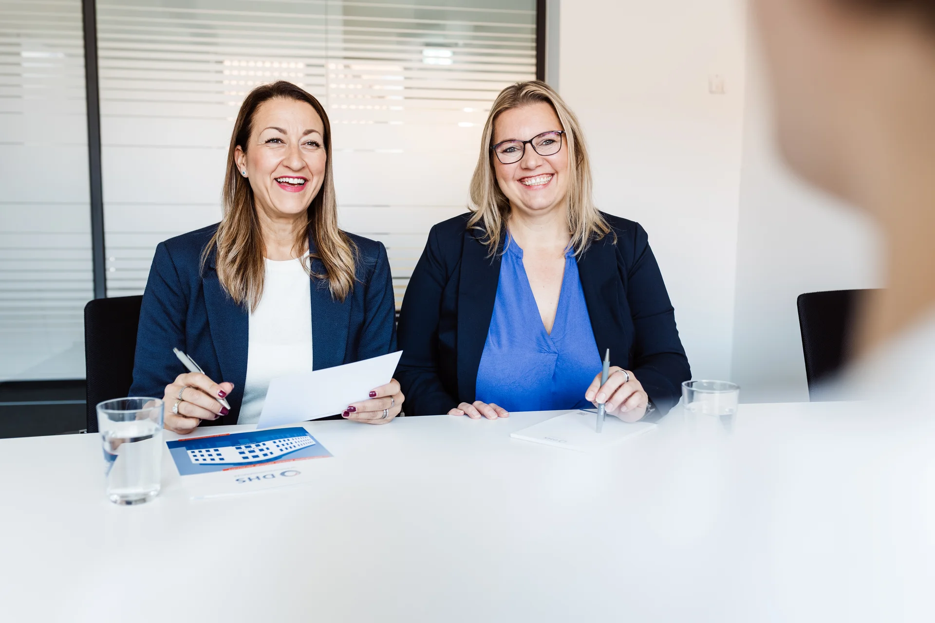 zwei Frauen in Buisinesskleidung sitzen am Tisch und lächerln in die kamera