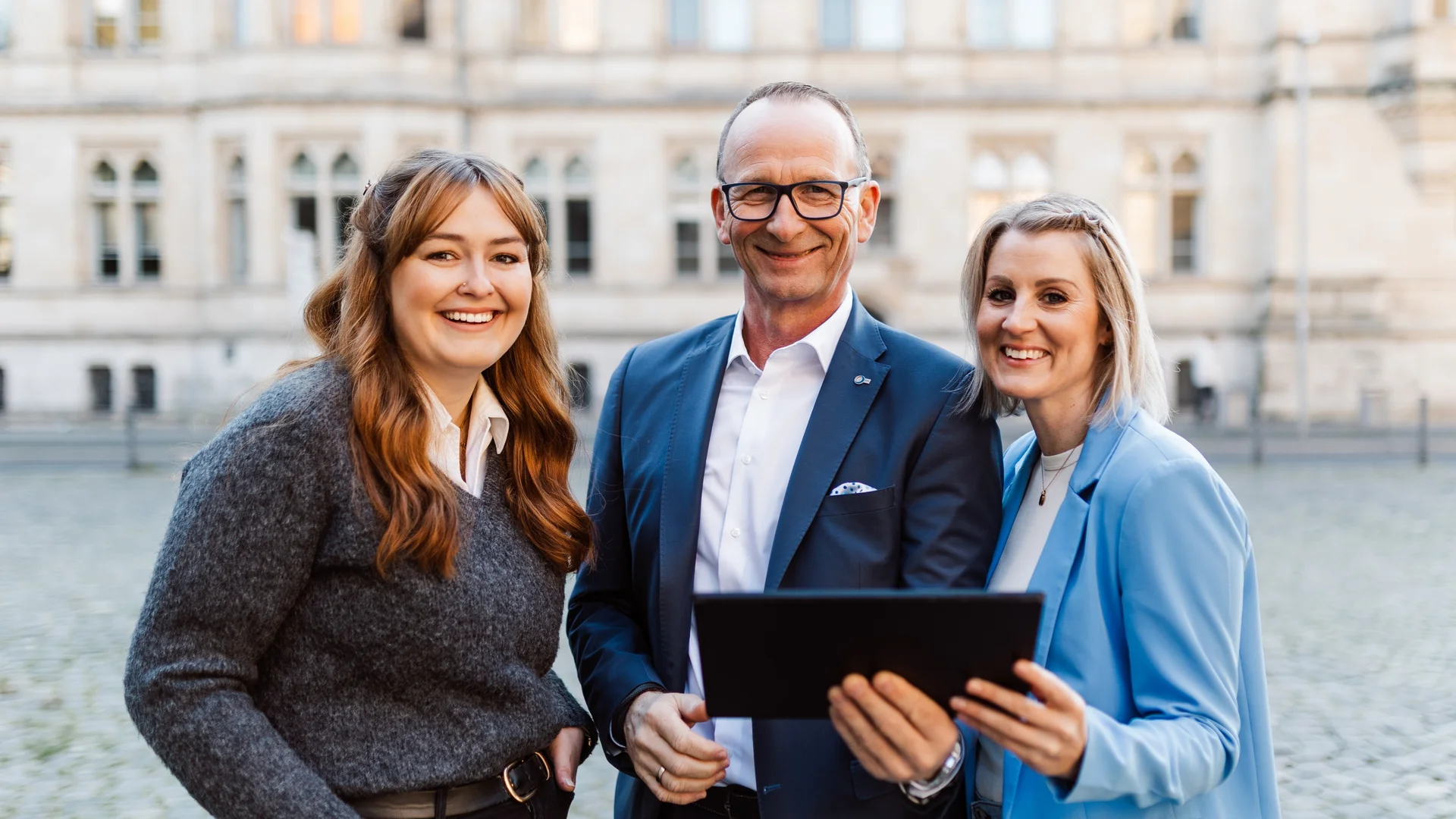 Eine Gruppe von drei Personen in Businesskleidung steht zusammen, der Mann in der Mitte hält ein Tablet in der Hand.