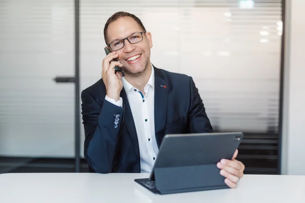 Lächelnder Mann mit Brille im Anzug telefoniert und hält Tablet in einem modernen Büro