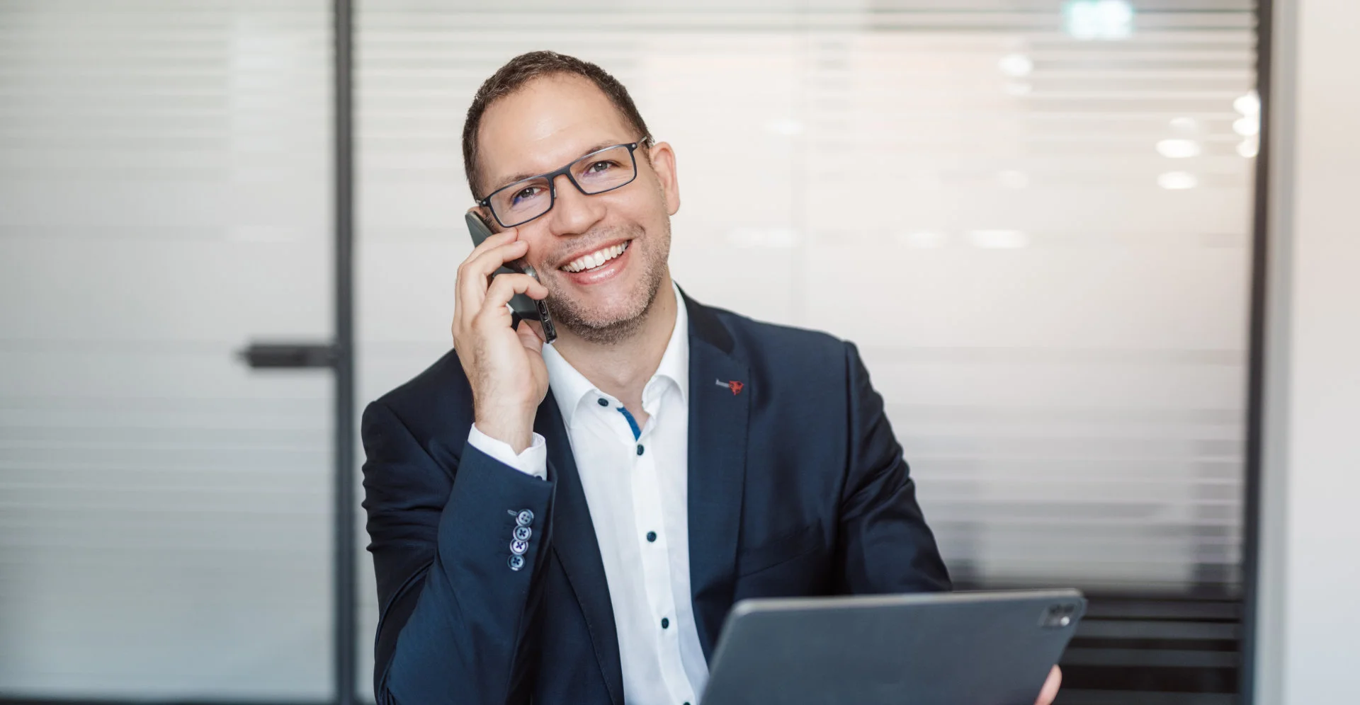 Lächelnder Mann mit Brille im Anzug telefoniert und hält Tablet in einem modernen Büro