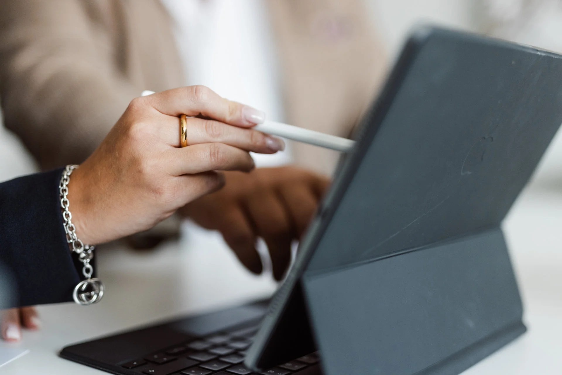 Hand mit goldfarbenem Ring und Stift zeigt auf Tablet-Bildschirm, weitere Hand tippt auf Tastatur