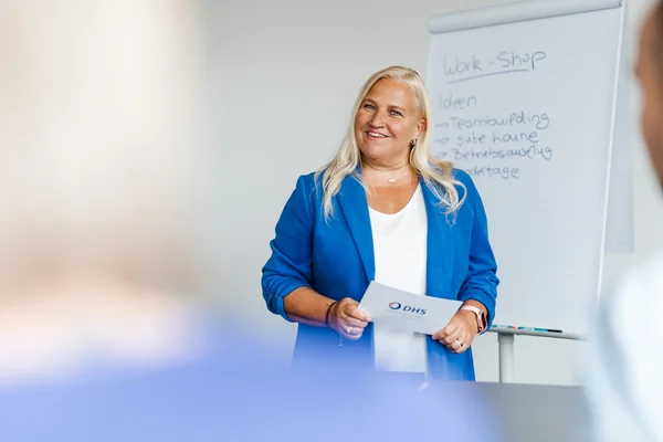 Blonde Frau in blauem Blazer steht neben Flipchart und lächelt in Kamera