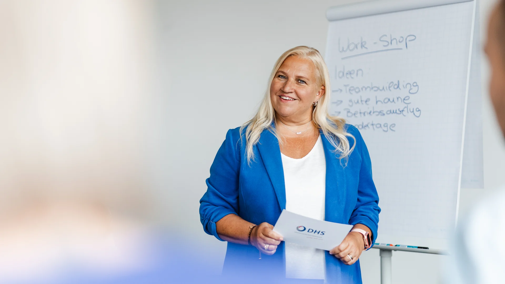 Blonde Frau in blauem Blazer steht neben Flipchart und lächelt in Kamera
