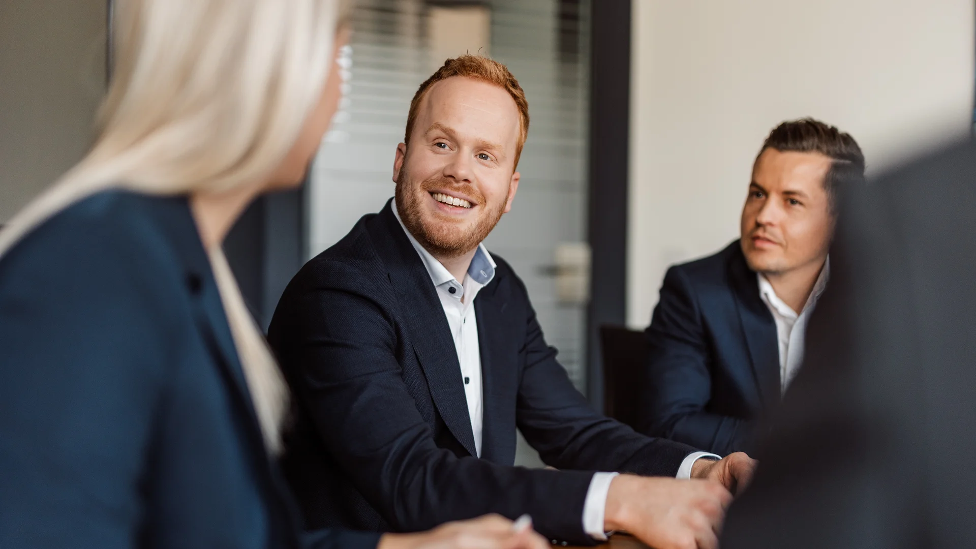 Es sind drei Personen in einer Besprechung und in Businesskleidung zu sehen, der Hinterkopf einer Frau, ein Mann der sie anlächelt und ein Mann der seitloch sitzt und ebenfalls lächelt