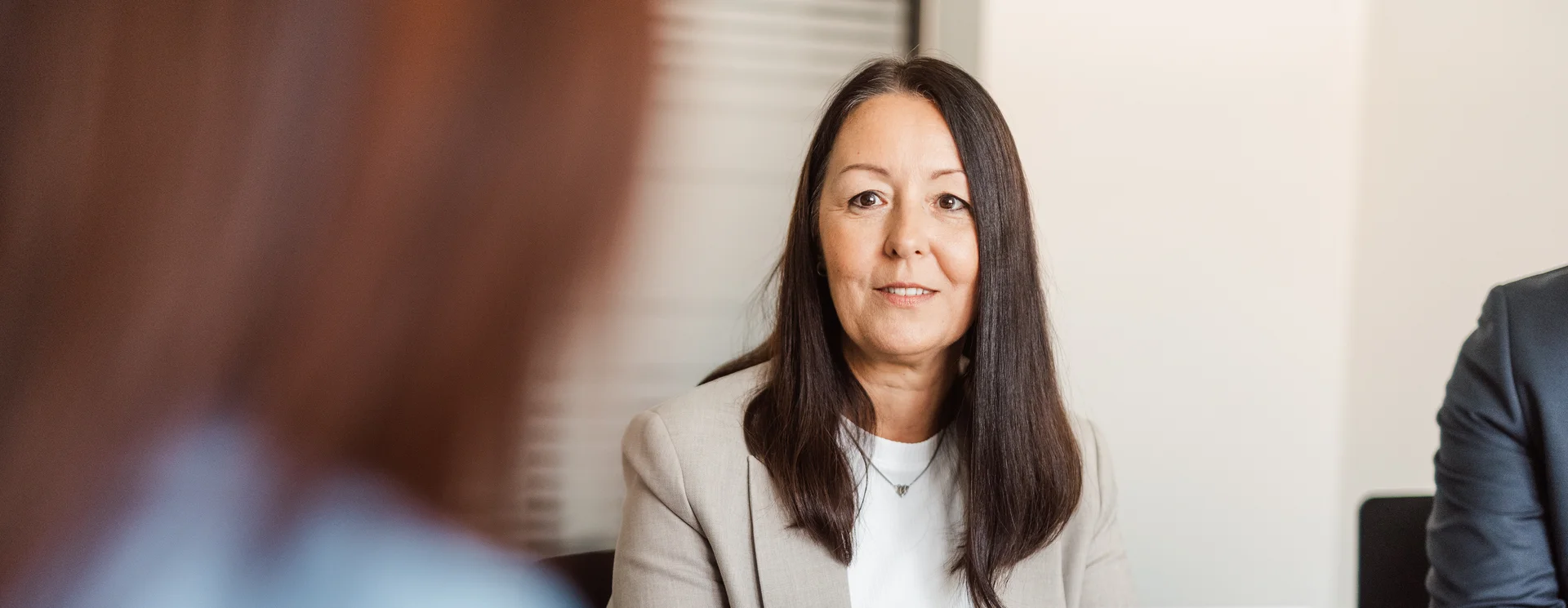 Eine Frau mit langen, dunklen Haaren trägt einen hellen Blazer und hält ein Tablet in der Hand. Ihr gegenüber sitzt eine weitere Person, von der man nur Hinterkopf und Schulter sieht.